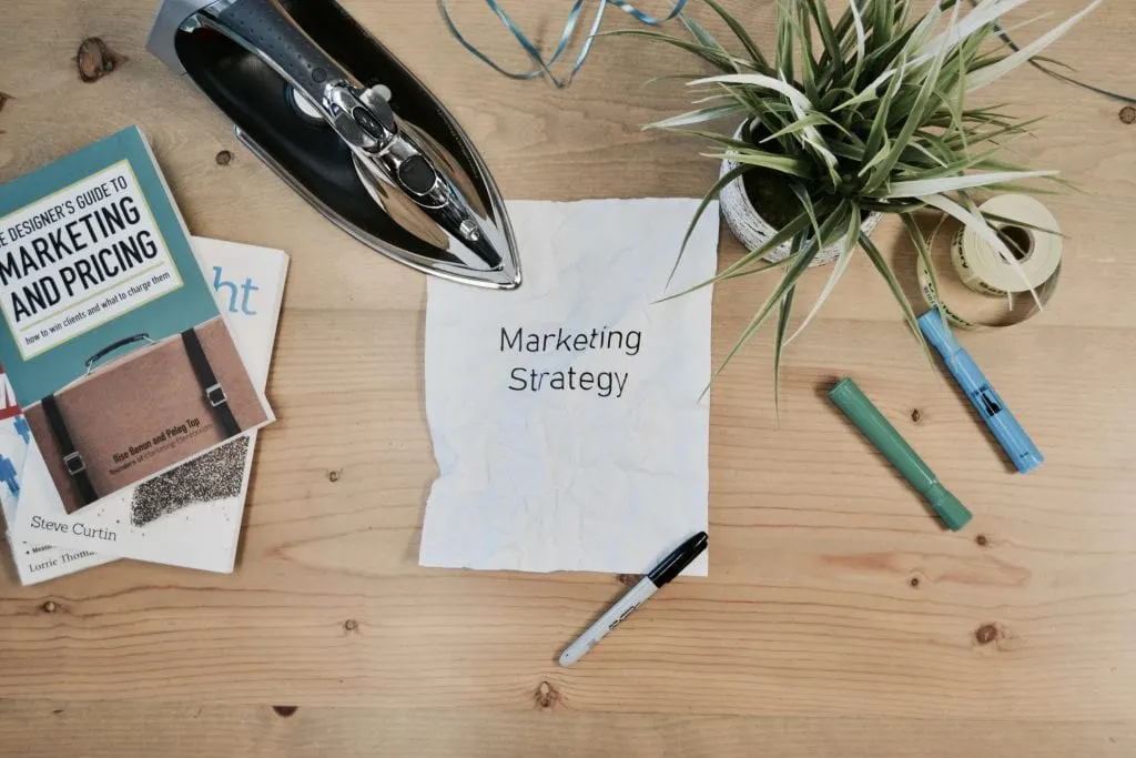 Marketing Strategy written on a paper on a wooden desk held down my a stapler, and plant. www.EverythingAbode.com Affiliate Marketing