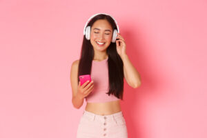 woman listening to podcasts with bright pink background smiling holding phone and wearing white headphones, fun indoor winter hobbies listening to podcasts