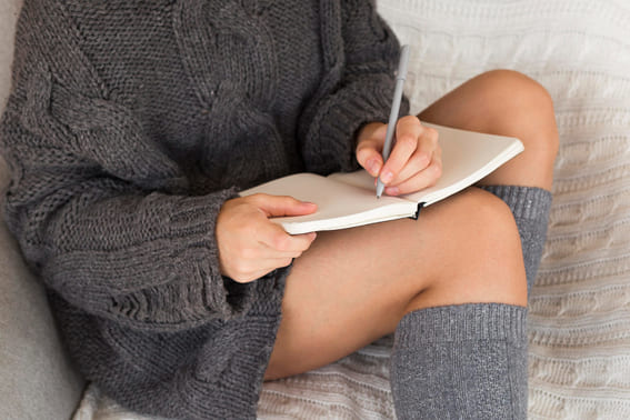 woman all curled up on bed with her grey socks and sweater writing in her journal as her self care routine