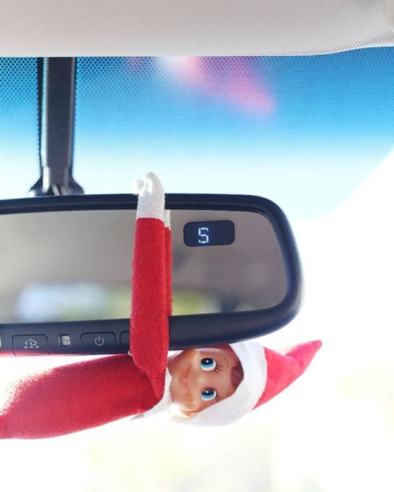 Elf on the Shelf dangles playfully from a car rearview mirror.