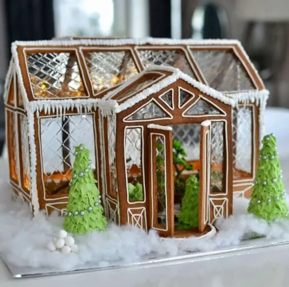 A gingerbread greenhouse with transparent windows and a glowing interior surrounded by icing trees.