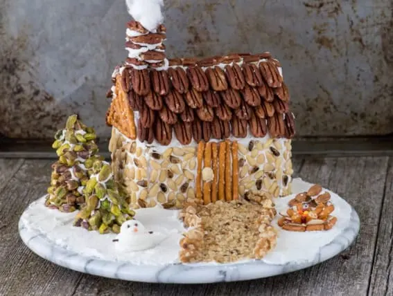 A gingerbread house adorned with almond shingles and a pecan log roof nestled in a snowy scene.