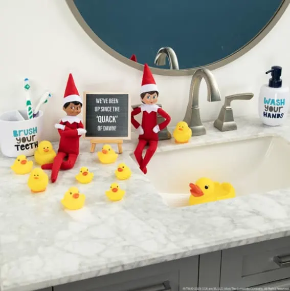 Elves on the Shelf seated among rubber ducks with a playful morning sign by the bathroom sink.