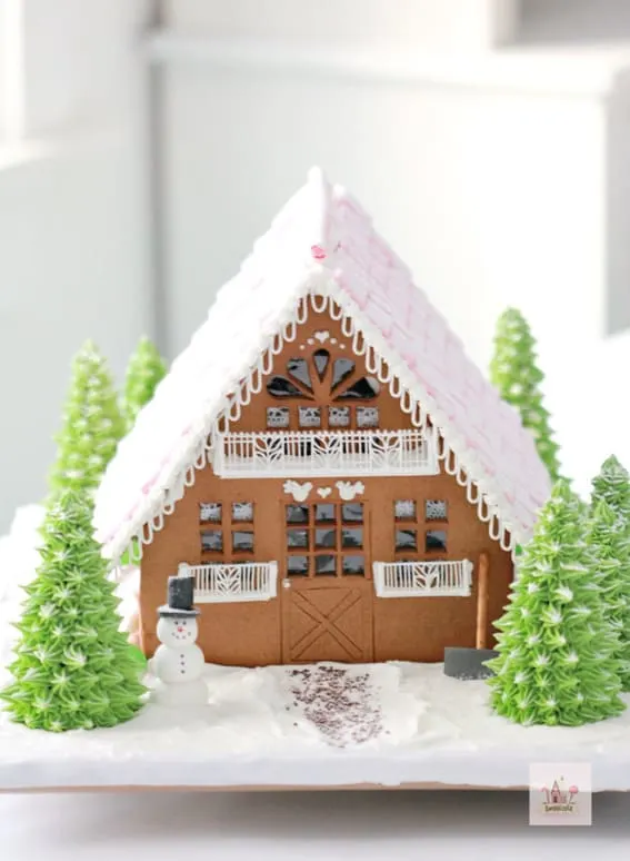 A charming gingerbread house with a pink icing roof and delicate white details flanked by green icing trees.