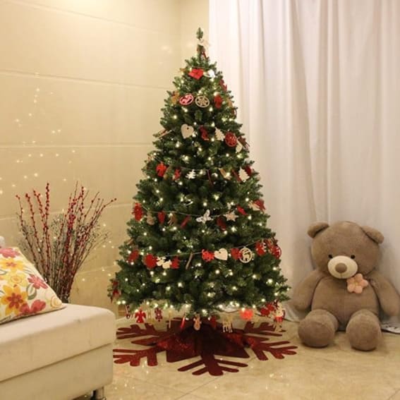 A 6-foot artificial pre-lit Christmas tree with a decoration skirt, 300 warm white LED lights, and adorned with various ornaments, beside a plush teddy bear.