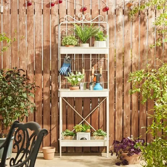 White metal garden shelf with plants and gardening tools against a wooden fence.