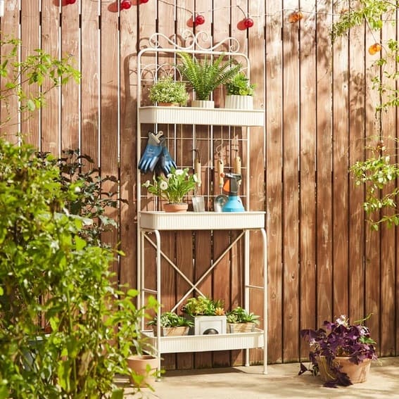 White metal garden shelf with plants and gardening tools against a wooden fence.