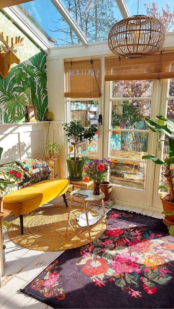 A sunny bohemian-style room with a yellow chair, a rattan table, and a colorful floral rug surrounded by indoor plants and bamboo window shades.