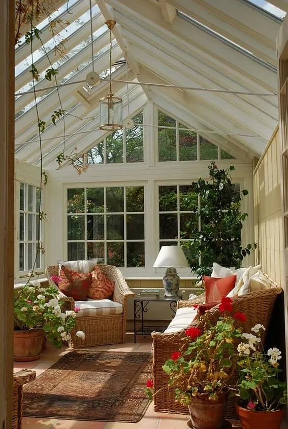 A bright conservatory with vaulted glass ceiling, wicker furniture, blooming plants, and terracotta flooring.