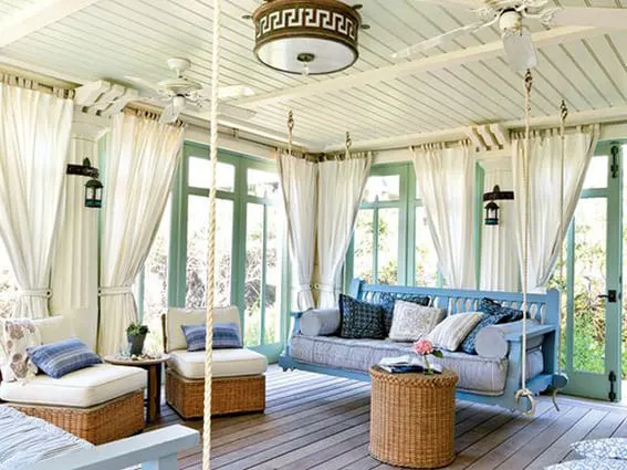 A bright sunroom with white drapes, a swing sofa, and wicker furniture on a wooden floor.