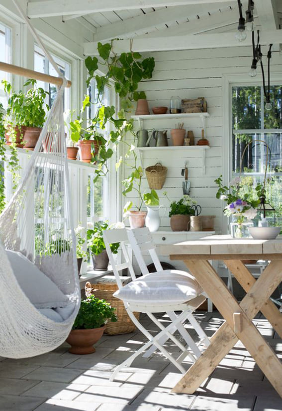 A fresh and airy sunroom with a white hammock, rustic wooden table, and shelves adorned with potted plants.