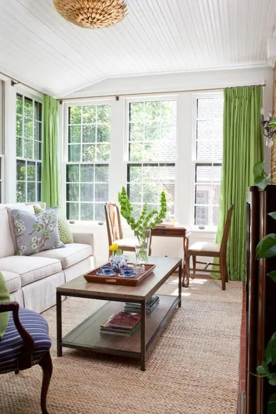 A classic sunroom with a plush sofa, wooden furniture, and green drapes against a backdrop of large windows.