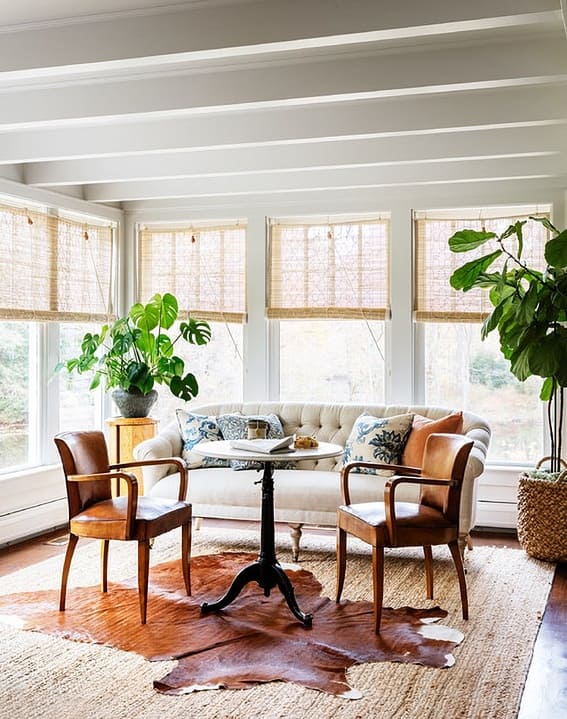 A serene sunroom with a comfy leather sofa, a beige couch and natural bamboo shades and a rug with a hide.
