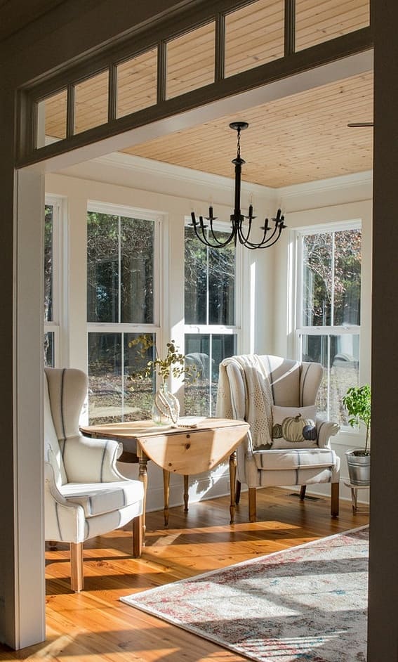 A sunroom bathed in golden light featuring elegant wingback chairs, a wooden table, and a classic chandelier.