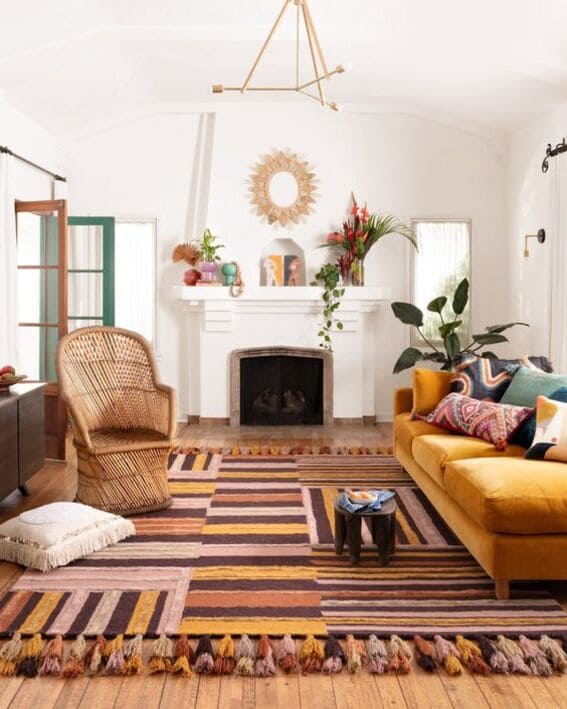A bright and airy living room with a bohemian flair, featuring a mustard yellow sofa, a large rattan armchair, colorful striped rugs with tassels, and a white fireplace flanked by green windows.
