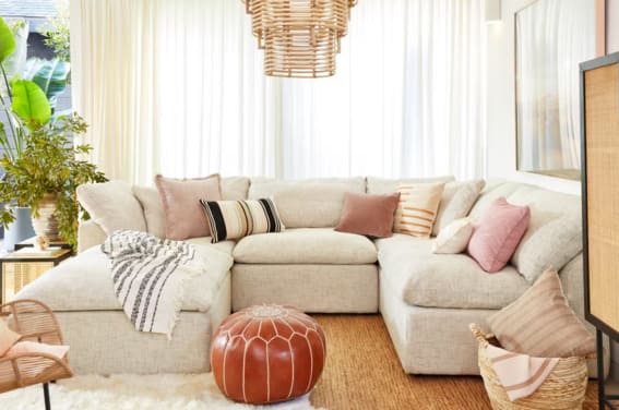A bright and inviting living room featuring a large U-shaped sofa covered with various textured pillows in shades of pink and beige, under a woven chandelier, with sheer curtains and lush plants enhancing the airy feel.