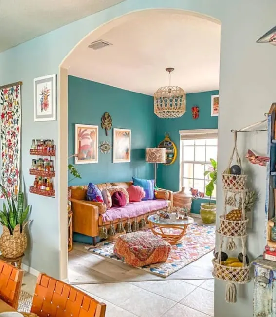 This vibrant and eclectic living room showcases bohemian decor, including a teal-blue wall, macramé wall hangings, and a colorful rug. The space also includes a leather sofa with pink cushions, a macramé ottoman, and a wicker chandelier.