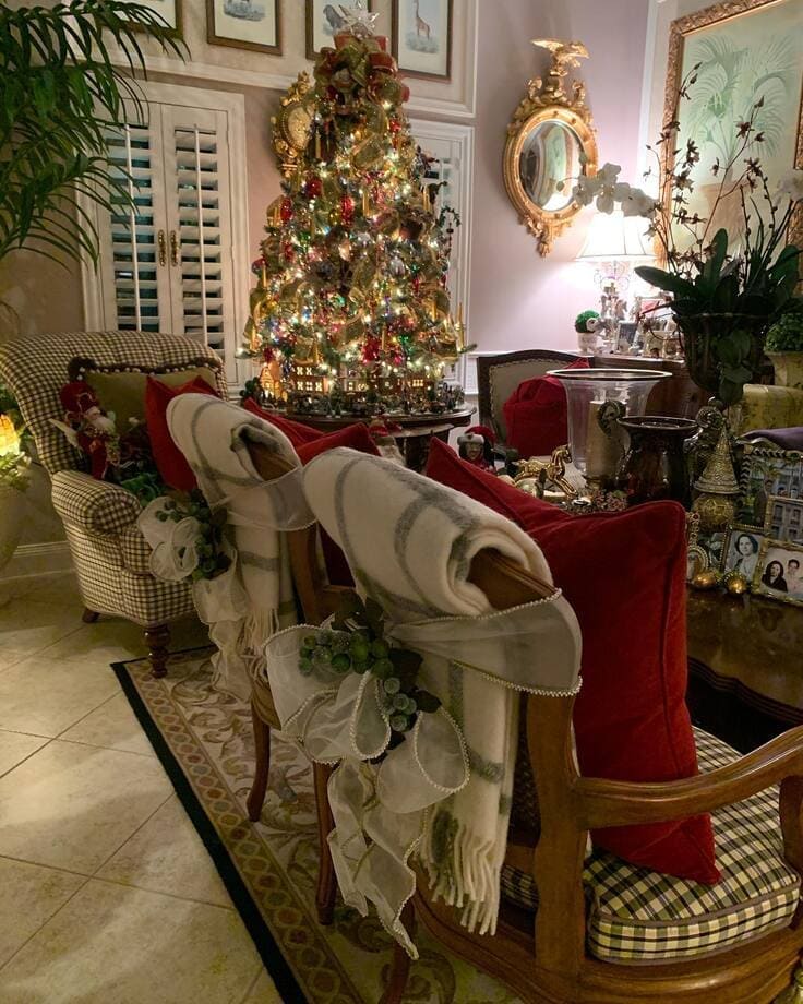 Ralph Lauren Christmas living room—ornate tree with gold ribbon, chair backs draped in ivory throws tied with organza and greenery, red velvet pillows, gingham seats, gilded mirror.