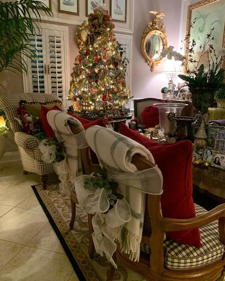 Ralph Lauren Christmas living room—ornate tree with gold ribbon, chair backs draped in ivory throws tied with organza and greenery, red velvet pillows, gingham seats, gilded mirror.