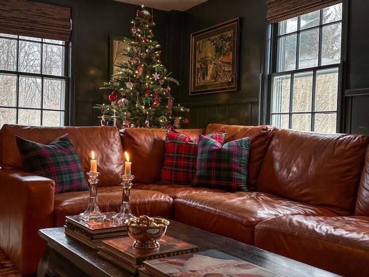 Dark-paneled Ralph Lauren Christmas lounge—caramel leather sectional, tartan plaid pillows, slim tree with classic ornaments, crystal candlesticks on stacked books.