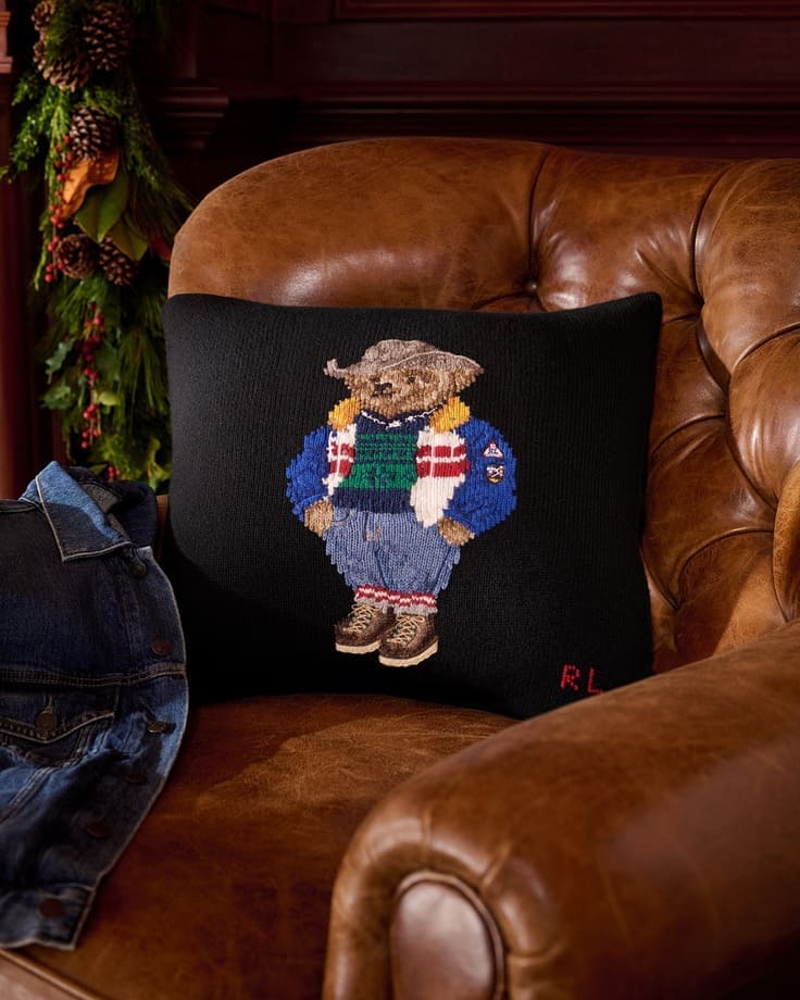 Ralph Lauren Polo Bear needlepoint pillow on a caramel leather club chair with pine garland—cozy Christmas vignette.