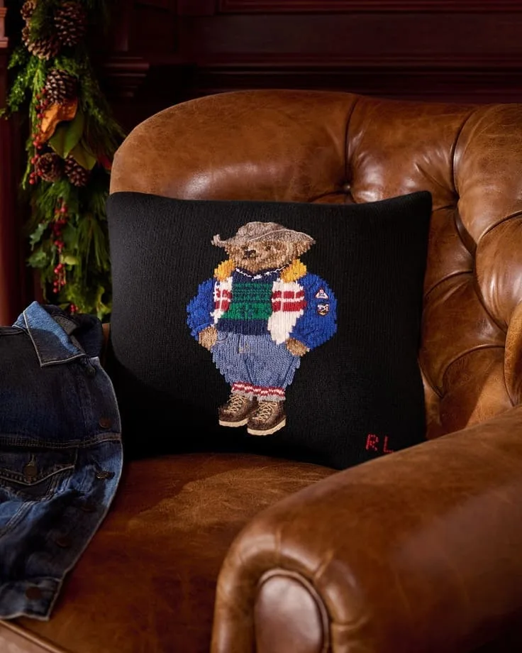 Ralph Lauren Polo Bear needlepoint pillow on a caramel leather club chair with pine garland—cozy Christmas vignette.