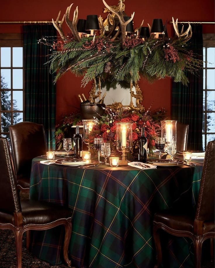 Ralph Lauren Christmas dining room—Black Watch tartan tablecloth, leather nailhead chairs, antler chandelier wrapped in greens, hurricanes, red florals, brass mirror.