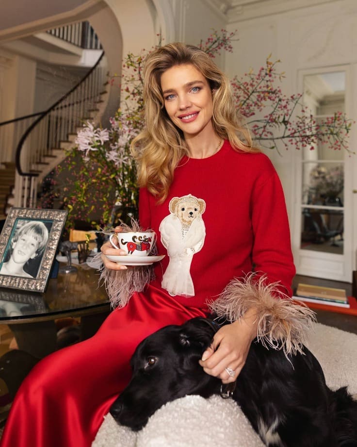 Ralph Lauren holiday moment—woman in red Polo Bear sweater with feather cuffs and satin skirt, holding a teacup, black dog at her lap, grand staircase behind.