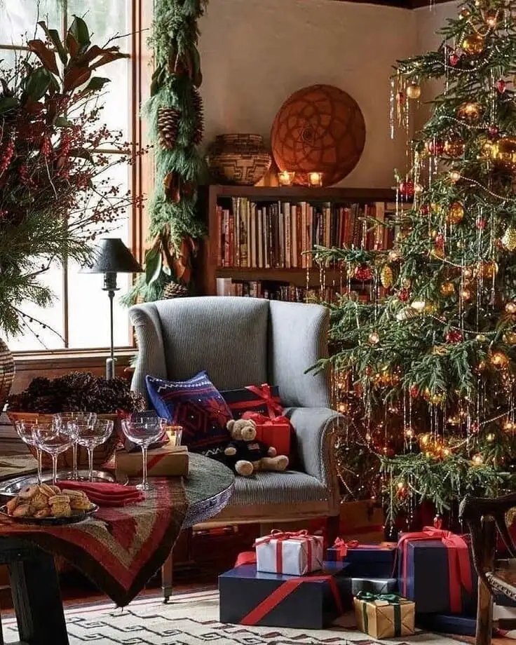 Ralph Lauren Christmas library—pinecone garland at the window, wingback chair with Southwestern pillow, tinsel-draped tree, navy-and-red wrapped gifts, teddy bear.