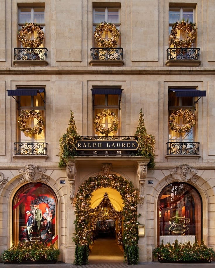 Ralph Lauren flagship facade with golden wreaths on iron balconies, garland-draped archway, navy awnings, glowing holiday windows. 