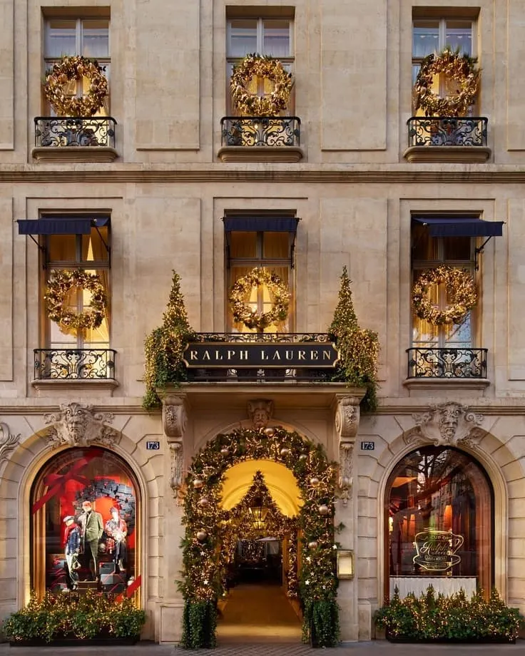 Ralph Lauren flagship facade with golden wreaths on iron balconies, garland-draped archway, navy awnings, glowing holiday windows.