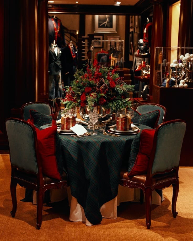 Tartan-clad holiday table—velvet chairs with red pillows, crystal stemware, evergreen-and-rose centerpiece, gift boxes at each place, dark wood clubroom vibe.