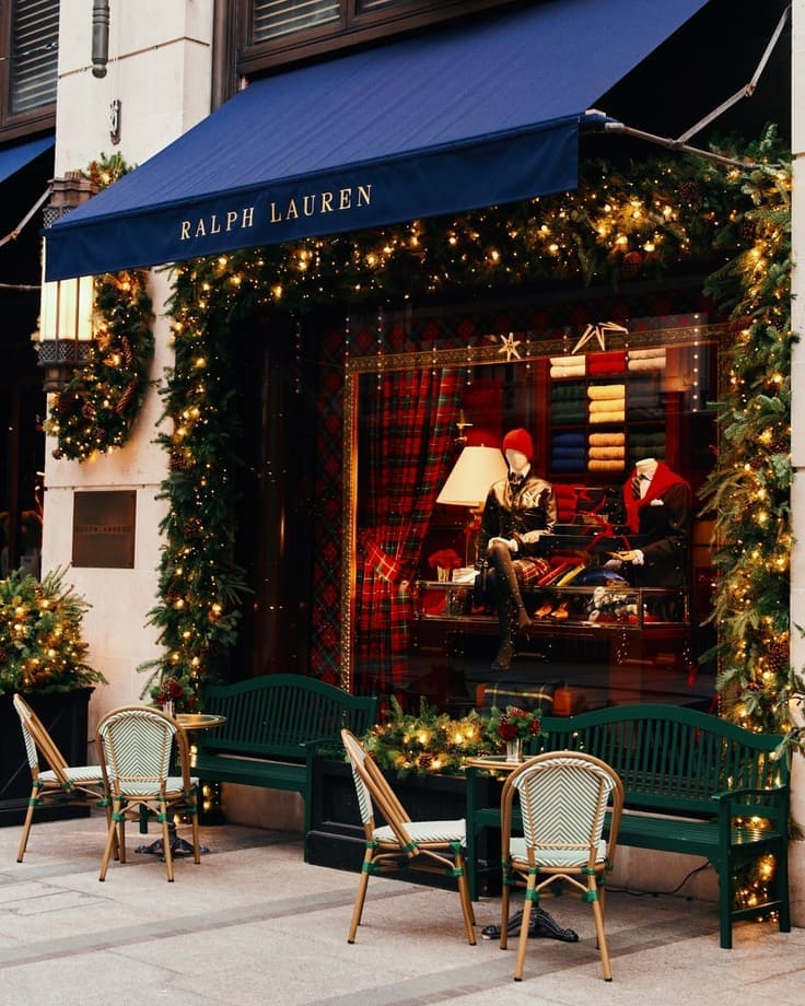 Ralph Lauren holiday storefront—navy awning, garland-wrapped windows with tartan backdrop, green bistro benches and café chairs, warm twinkle lights.