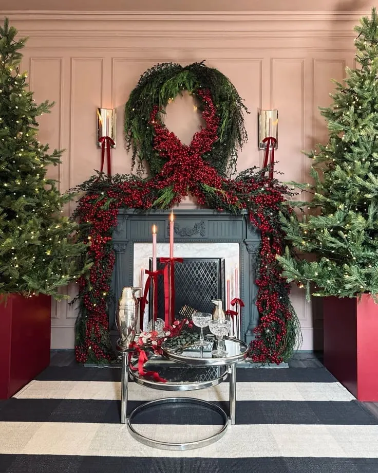 Ralph Lauren–style fireplace—oversized red-berry garland with bow, charcoal mantel, blush paneled walls, twin trees in red planters, striped rug, crystal bar setup with red tapers.
