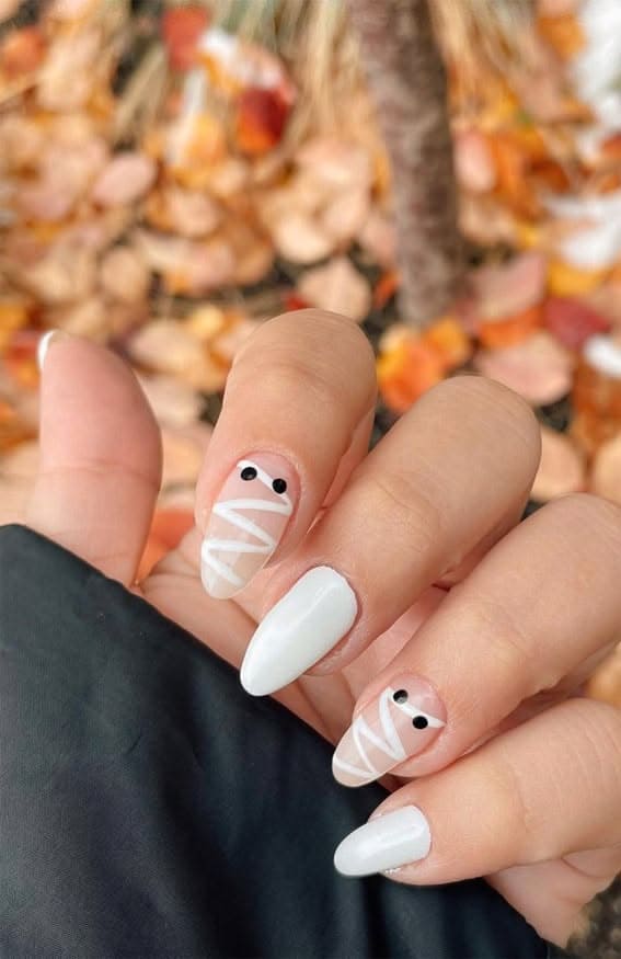 Simple white Halloween nails with mummy accent nails and black dot eyes on almond shaped nails