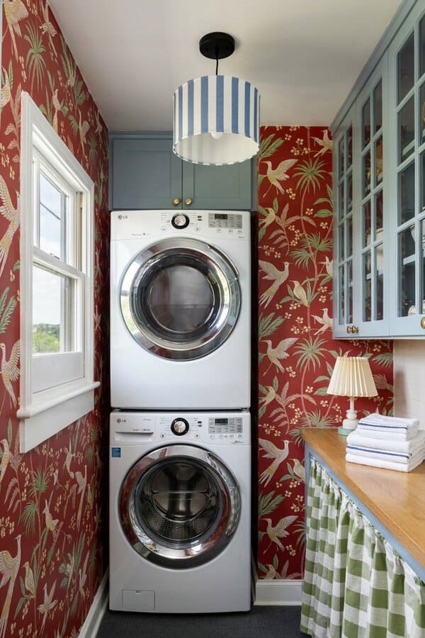 Narrow laundry room with a stacked washer and dryer, red botanical wallpaper and blue cabinetry.